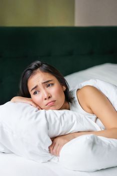 Sad and gloomy asian girl lying in her bed, grimacing and crying, feeling upset, spending time in her bedroom, hugging pillow.