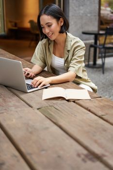 Portrait of stylish urban woman working on laptop from outdoor cafe, sitting with documents and computer, smiling.