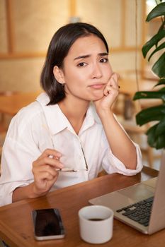 Vertical shot of sad and gloomy young woman, sitting with laptop in cafe, grimacing and looking upset, feeling tired or disappointed.