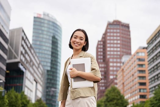 Portrait of young asian woman, looking happy and confident, going to work or university, city skyscrappers behind her, holding laptop and notebook.