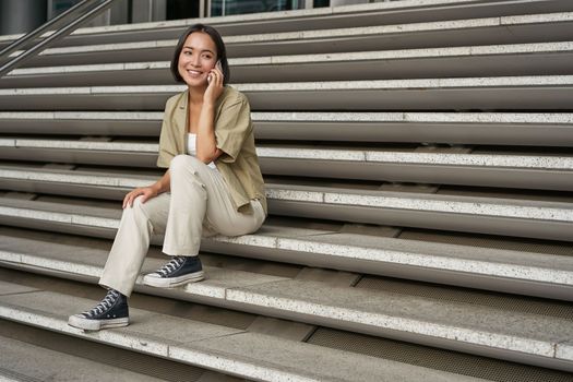 Smiling asian girl sits on stairs of building and talks on mobile phone, relaxing during telephone conversation.