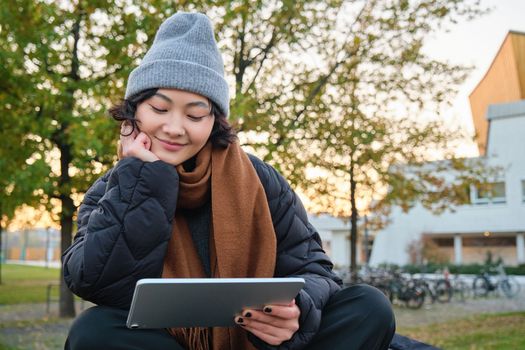 Smiling asian girl, student sits on bench in park alone, reading, using social media app on digital tablet, watches videos outdoors, relaxes on fresh air.