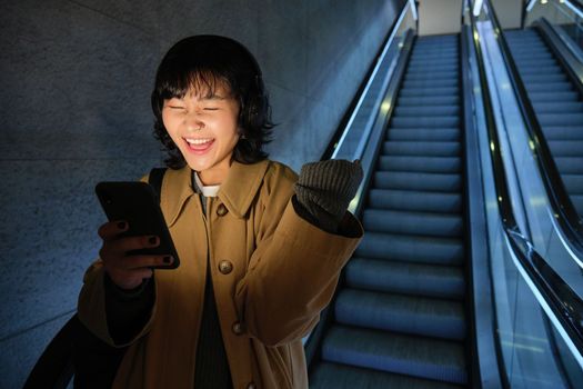 Urban lifestyle and people. Portrait of girl feeling happy and excited, going down escalator in headphones, looking at smartphone and celebrating.