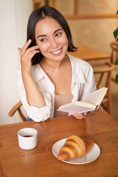 Vertical shot of happy young asian woman enjoys reading, sitting with book in cafe, drinking coffee and eating croissant.