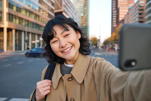 Portrait of young asian woman taking selfie in front of building in city centre, tourist takes photos while sightseeing, smiling at smartphone camera.