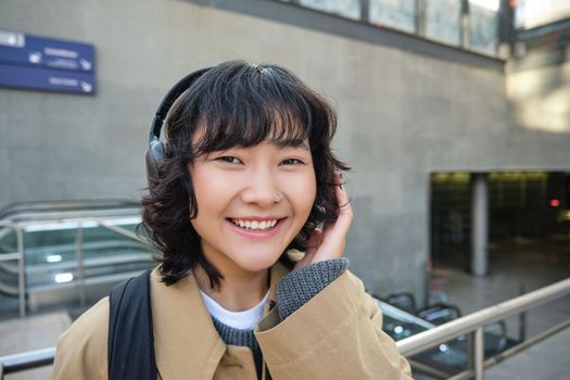 Close up of smiling brunette girl in headphones, listens music, travels around city, commutes to work, stands on a street.