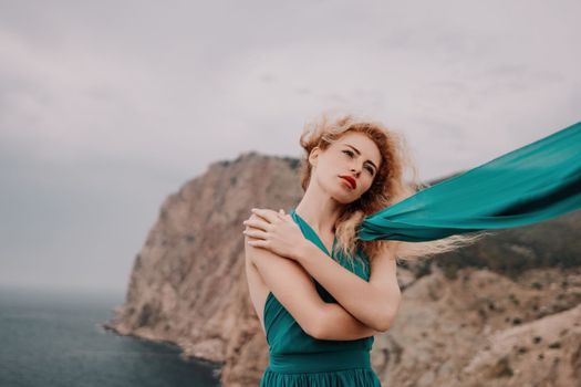 Side view a Young beautiful sensual woman in a mint long dress posing on a volcanic rock high above the sea during sunset. Girl on the nature on overcast sky background. Fashion photo