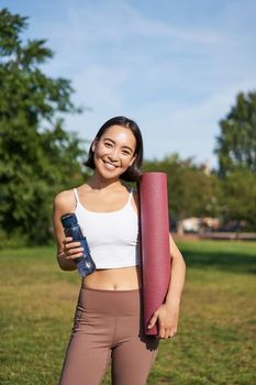 Portrait of young slim and healthy korean girl doing workout in park, standing with water bottle and rubber mat for execises on green lawn, smiling happily.