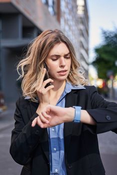 Vertical portrait of businesswoman having a phone call, looking at time on her digital watch, reading message on device.