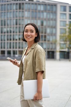 Vertical shot of asian girl walks with laptop on city street, smiles.