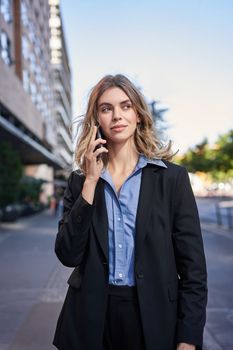 Vertical shot of confident young businesswoman making phone call, standing on street and talking on telephone.