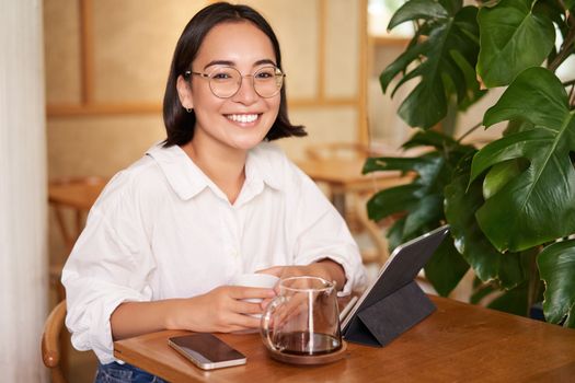 Smiling asian girl in glasses, woman working on remote, drinking coffee and using tablet. Copy space
