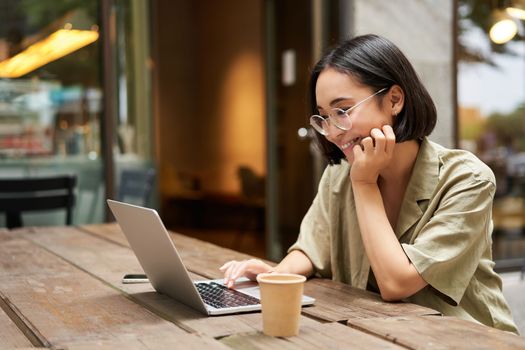 Portrait of smiling girl in glasses, sitting with laptop in outdoor cafe, drinking coffee and working remotely, studying online.