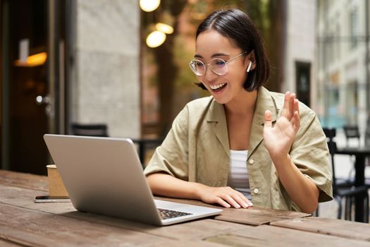 Portrait of young woman video chatting, having online meeting on laptop, sitting in outdoor cafe with compute, wearing glasses and smiling.