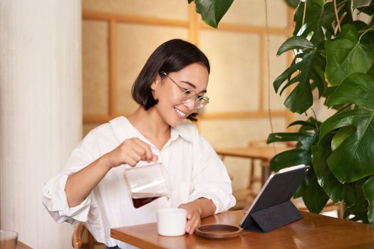 Freelance and remote workers. Smiling young woman pouring coffee in a cup, sitting in cafe and looking at digital tablet.