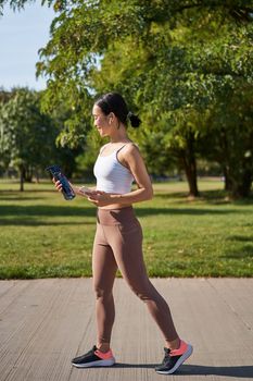 Active asian girl, in fitness clothing, workout in park, walking in sportswear with smartphone and water bottle.