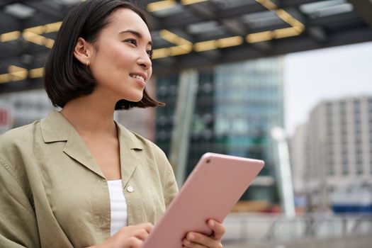 Portrait of asian woman standing on street, using tablet, smiling with carefree face expression, outdoor shot.