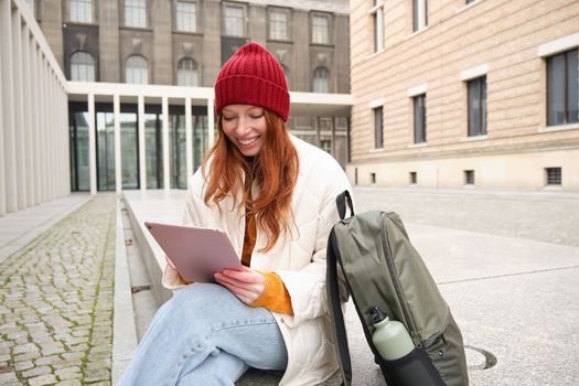 Redhead girl smiles, sits outdoors near building with digital tablet, thermos and backpack, connects to public internet and searches smth online on her gadget.