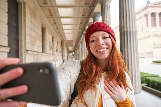 Cute young redhead woman takes selfie on street with mobile phone, makes a photo of herself with smartphone app on street.