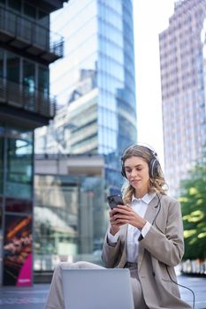 Corporate woman in suit, sitting in city, listening to music with laptop, scrolling news on her mobile phone, relaxing on lunch break.