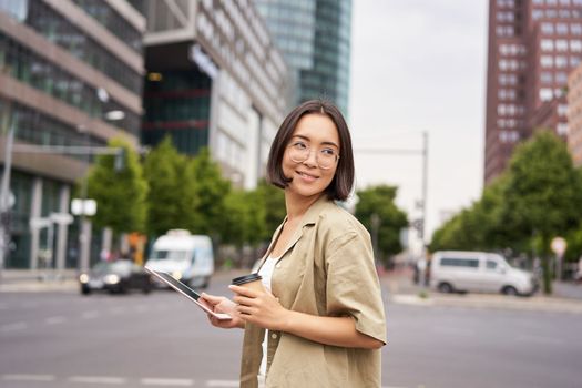 Happy woman exploring the city. Young korean girl holds tablet, drinks coffee and walks along street with big smile on her face.