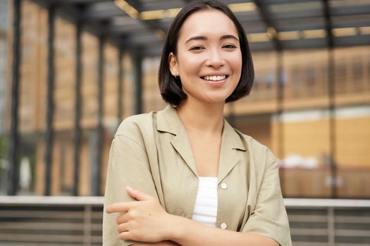 Portrait of young confident female model, girl in casual clothes, posing outside near glass building, smiling at camera.