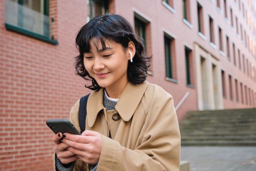 Happy korean girl walks on street, listens music in wireless earphones and holds smartphone, picks song in playlist while standing outdoor near building, reading message.