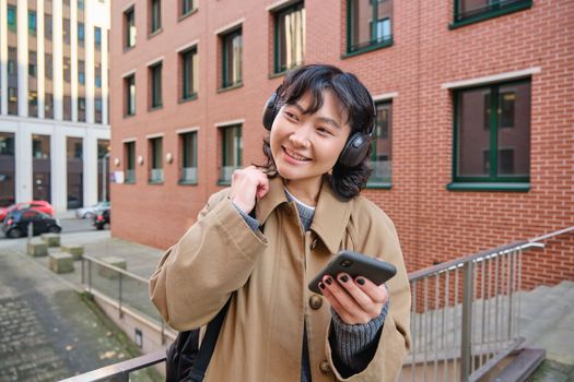 Young happy woman in trench coat, listens music in headphones, holds smartphone, uses mobile application to find route in new city, stands on street with backpack.