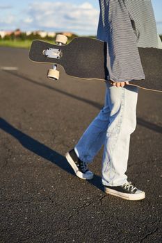 Cropped shot of teen skater girl, hands holding longboard, walking with skateboard on concrete road.