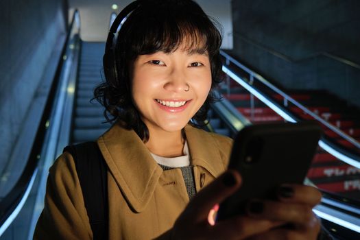 Close up of cute asian girl in headphones, picks song to listen while commuting, going down escalator to metro, smiling as reading message on mobile phone.