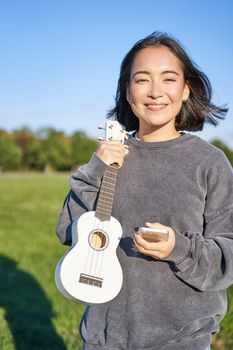 Vertical shot of young woman shows her ukulele, holds smartphone in hand and looks at camera. People and hobbies concept