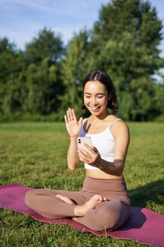 Portrait of asian girl sitting on mat in park, talking to smartphone, meditating online with yoga instructor on fresh air.