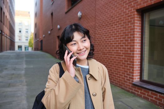 Portrait of young korean woman walking down street with phone, talking with someone, makes a call, has telephone conversation.