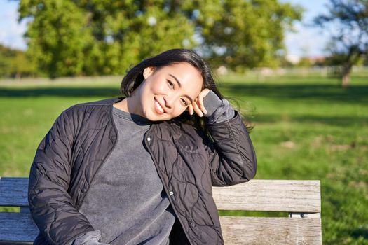 Beauty portrait of smiling asian girl with dark short hair, sitting on bench in park and gazing at camera.