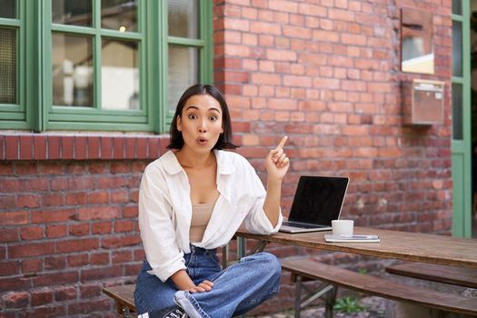 stylish modern asian girl with laptop, sitting in cafe, looking amazed and pointing at upper right corner banner, showing info advertisement.