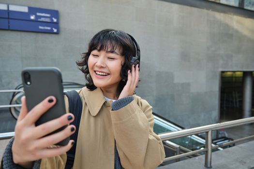 Beautiful korean girl in headphones, takes selfie on her smartphone, stands on street with mobile phone, records video, listens music and travels around city.