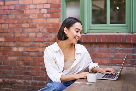 Stylish young businesswoman, asian girl with laptop, sitting in outdoor cafe with cup of coffee and working, using computer.