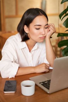 Vertical shot of asian girl looking tired at laptop screen, bored of working, sitting in cafe sleepy and exhausted.