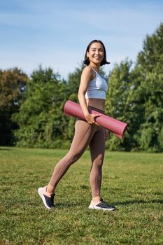 Vertical shot of stylish fitness woman with rubber mat, posing on lawn in park, doing workout on fresh air.