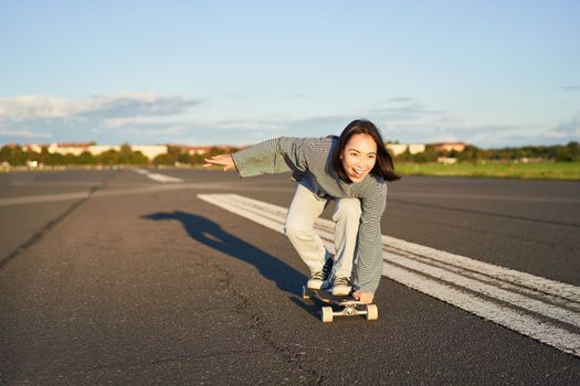 Carefree skater girl on her skateboard, riding longboard on an empty road, holding hands sideways and laughing.