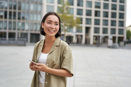 Portrait of asian woman standing on street, city square and holding mobile phone. Girl with smartphone walking outdoors.