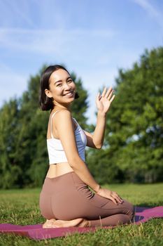 Portrait of asian girl on fitness class in park, sitting on rubber mat and wave hand at camera, say hello, meditating and practice yoga.