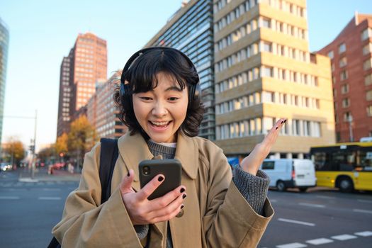 Young happy woman celebrating on street, holding smartphone and cheering, reacts amazed to good news, reads phone text message with surprised joyful face.