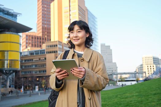 Happy young brunette girl, asian woman walks around city with tablet, goes to university with her digital gadget and backpack.