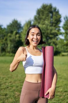 Beautiful and fit fitness girl in leggings, shows thumb up in park, stands with yoga rubber mat for workout and sport training.