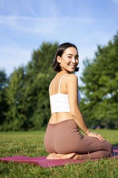 Portrait of asian woman sitting and meditating in park, doing yoga on fresh air, fitness exercises, smiling at camera.