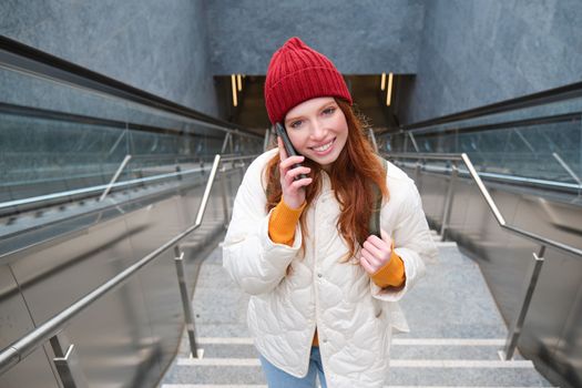 Portrait of happy redhead woman walking around town with smartphone, calling someone, talking on mobile phone outdoors.