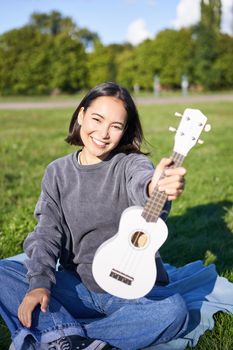 Music and instruments. Smiling asian girl shows her white ukulele, sits in park and plays small guitar.