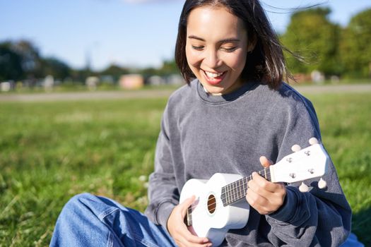 Portrait of asian girl student, playing ukulele and singing in park, sitting alone on blanket and enjoying making music.