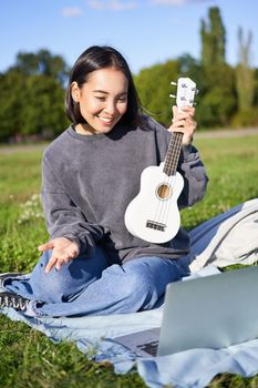 Vertical shot of happy asian girl plays instrument, shows her ukulele at laptop camera, video chats about music, teaches how to play, sits in park outdoors.